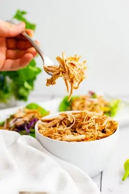 Whole30 slow cooker cajun chicken recipe shredded in a large white bowl with a fork filled with shredded jerk chicken above the bowl.