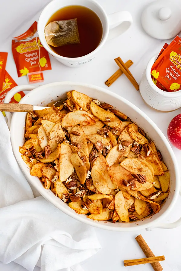 Large white oval casserole dish filled with healthy baked sliced apples with a wooden spoon in the apples with apples and Bigelow tea bags in the background.