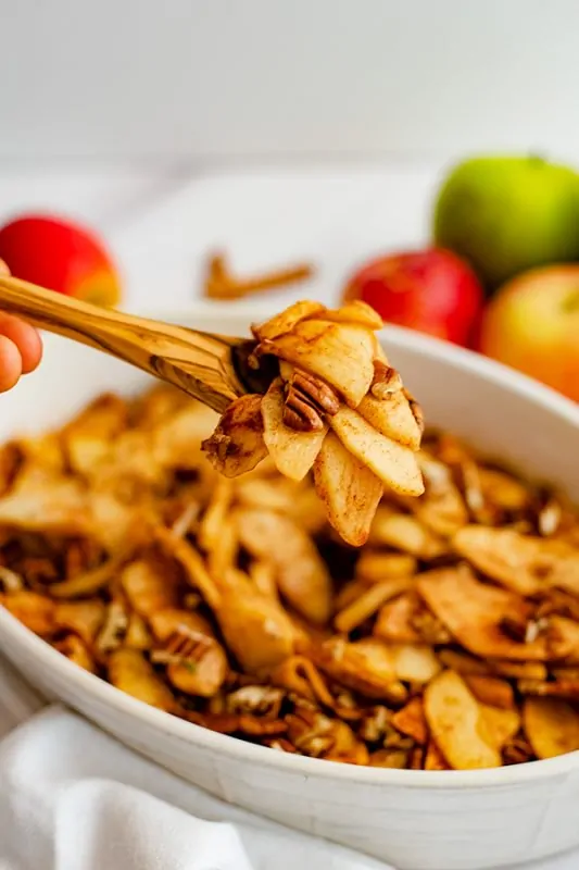 Wooden spoon holding a spoonful of healthy baked sliced apples over an oval white casserole dish filled with baked apples.