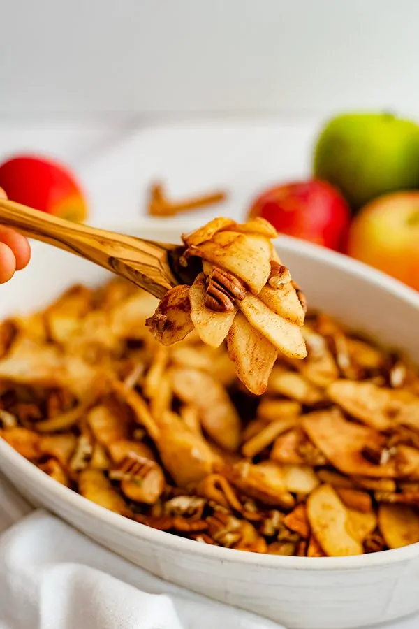 Wooden spoon holding a spoonful of healthy baked sliced apples over an oval white casserole dish filled with baked apples. 