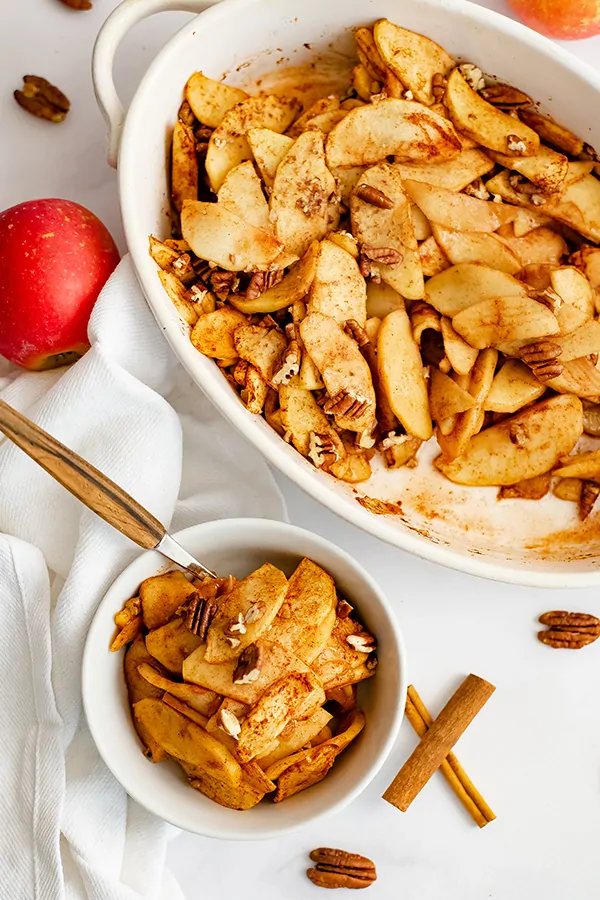 Small white bowl filled with healthy baked sliced apples with a white napkin and cinnamon sticks and pecans around the bowl. In the background is the casserole dish with some of the apples removed