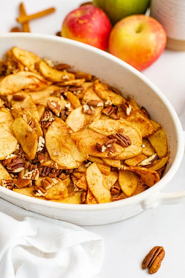 Large white oval casserole dish filled with healthy baked sliced apples surrounded by a white napkin, pecans, apples and cinnamon sticks. 