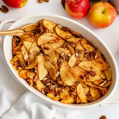 Large white oval casserole dish filled with healthy baked sliced apples with a wooden spoon in the apples with apples in the background.