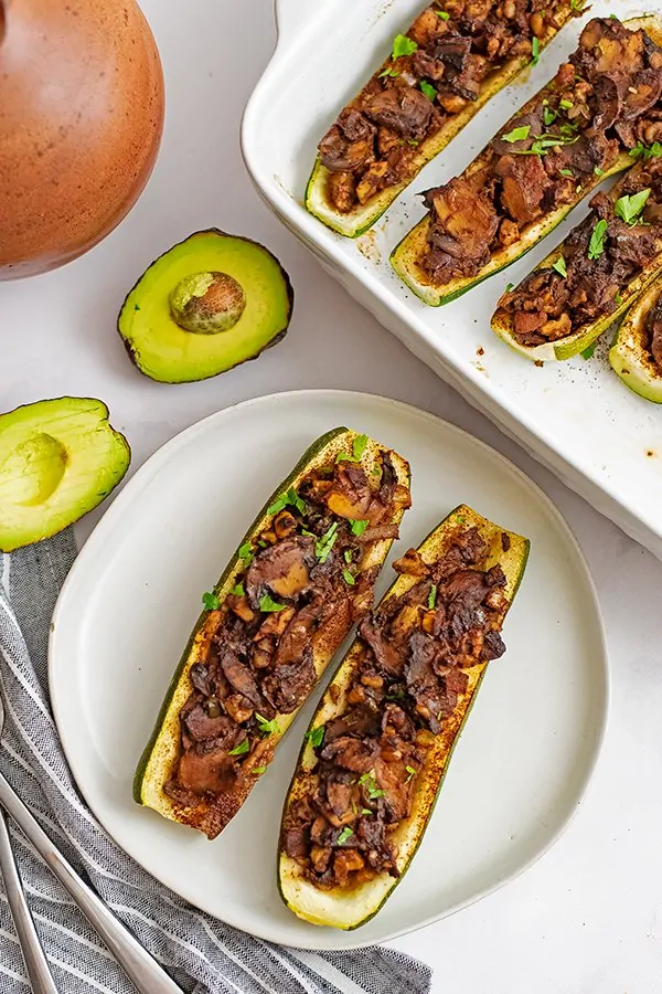 Overhead shot of Two Mexican Stuffed Zucchini Boat Recipe on 2 white plates stacked on top of each other with an avocado and a white ceramic dish filled with stuffed zucchini