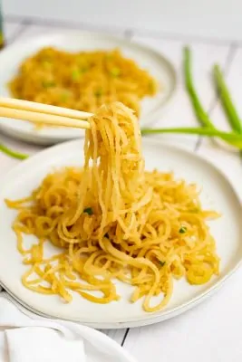 White plate filled with sesame Asian low carb noodles with chopsticks picking up the noodles, bringing the noodles into focus