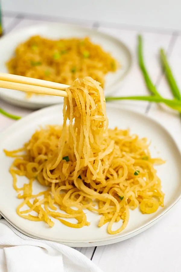 White plate filled with sesame Asian low carb noodles with chopsticks picking up the noodles, bringing the noodles into focus