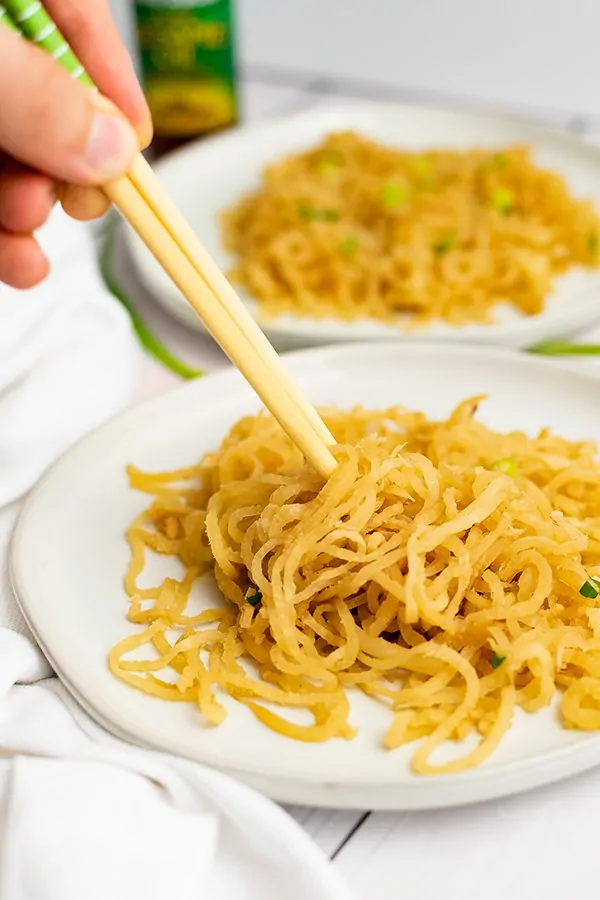 Chopsticks being held by a hand on the left side of the photo picking up sesame asian low carb noodles on a white plate with a white napkin on the left side of the plate. 