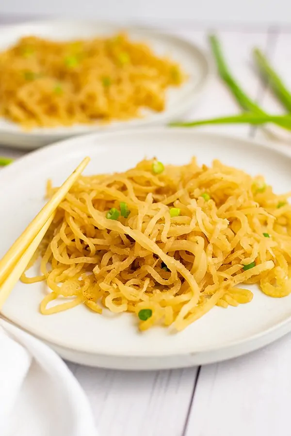 White plate filled with sesame asian low carb pasta with chopsticks sitting on the plate and a white napkin under the plate. There are green onions in the background