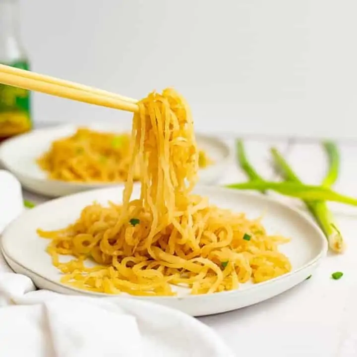 White plate filled with sesame Asian low carb noodles with chopsticks picking up the noodles, bringing the noodles into focus with green onions in the background