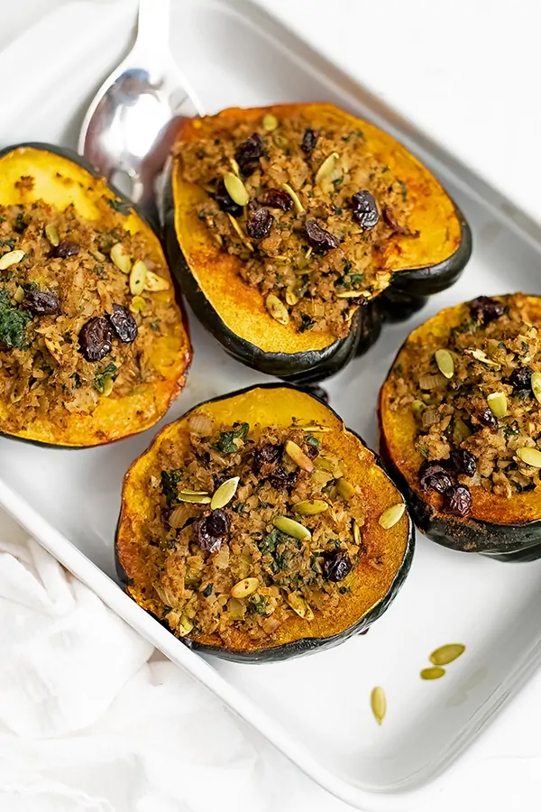 Ceramic white baking pan with 4 stuffed acorn squash in the pan with extra pumpkin seeds sprinkled around.