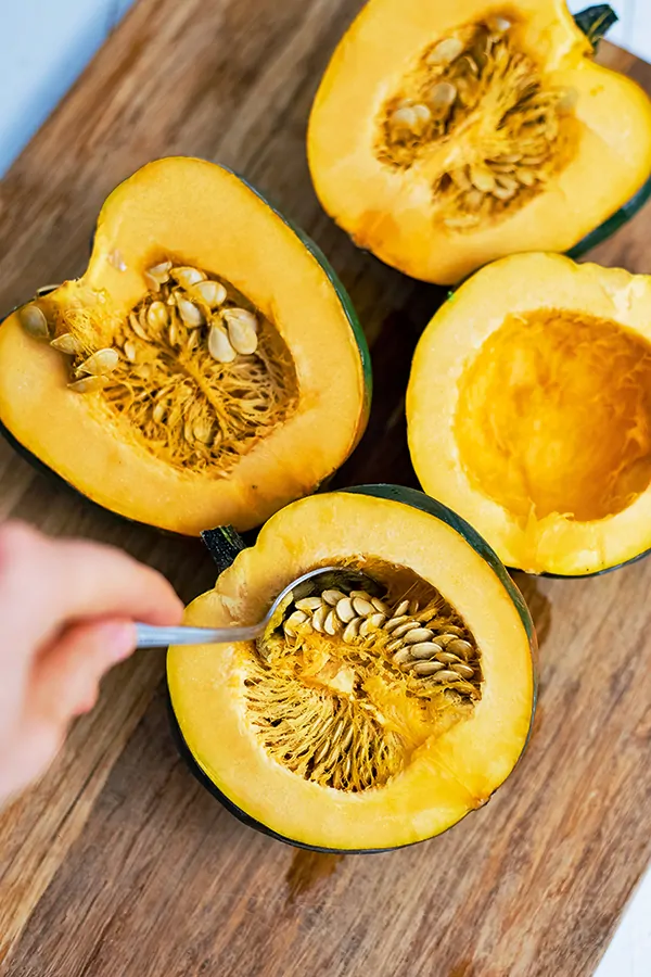 Brown cutting board with acorn squash seeds being scooped out with a spoon before becoming the stuffed acorn squash recipe.