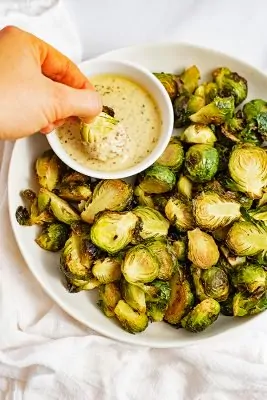 Large plate filled with crispy roasted brussel sprouts with garlic dijon sauce with a brussel being dipped into the sauce
