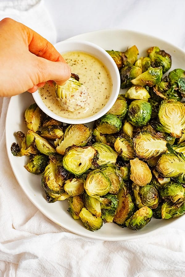 Large plate filled with crispy roasted brussel sprouts with garlic dijon sauce with a brussel being dipped into the sauce