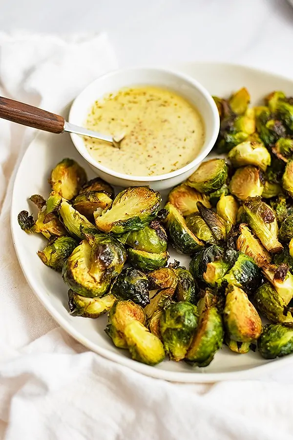 Overhead shot of a plate of crispy roasted brussel sprouts with a small bowl of garlic dijon sauce in the top left side of the bowl over a white napkin