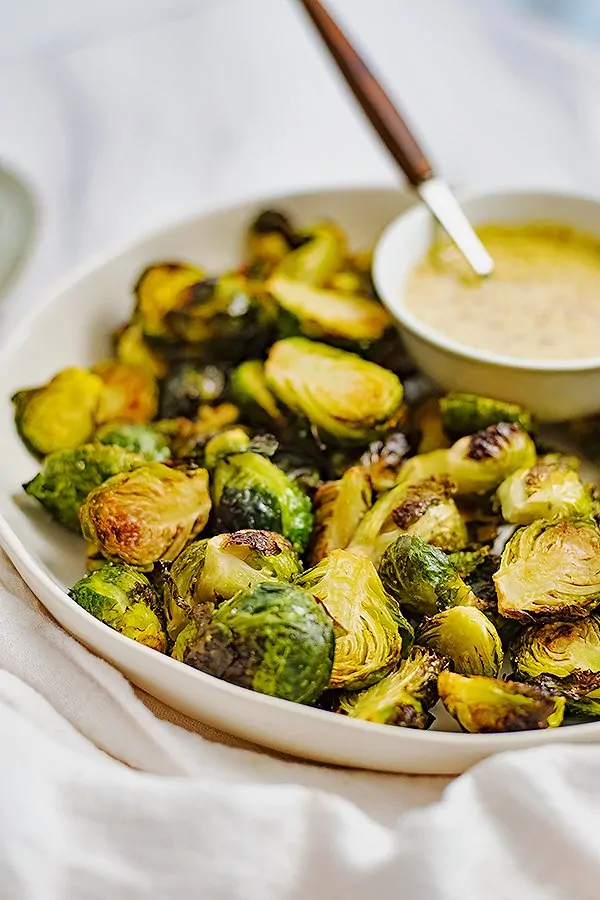 Large white plate filled with crispy roasted brussel sprouts with a small white bowl filled with garlic dijon sauce in the top right corner of the plate with a wooden spoon in the sauce