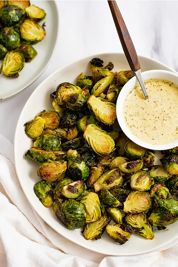 Overhead shot of Large white plate filled with crispy roasted brussel sprouts with a small white bowl filled with garlic dijon sauce in the top right corner of the plate with a wooden spoon in the sauce