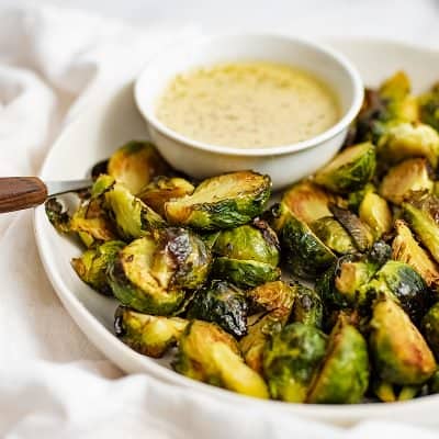 Crispy roasted brussel sprouts on a white plate with a bowl of garlic dijon sauce on a white napkin