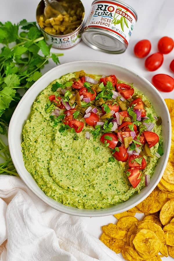 Large bowl filled with jalapeno cilantro hummus topped with tomatoes and red onion with cilantro, cherry tomatoes and plantains around the bowl.