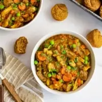 Bowl of easy beef stew (made in instant pot) with muffins around the bowl. Brown striped napkin with wooden spoons to the left