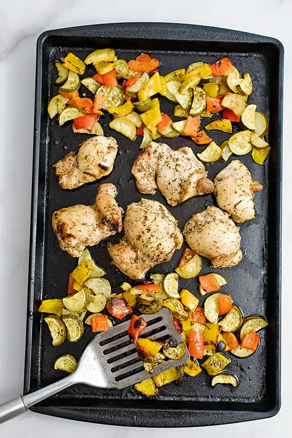Sheet pan filled with cooked greek chicken marinade sheet pan dinner with a silver spatula in the veggies