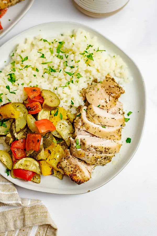 Overhead shot of a white plate filled with greek chicken marinade sheet pan dinner and cauliflower rice over a brown striped napkin