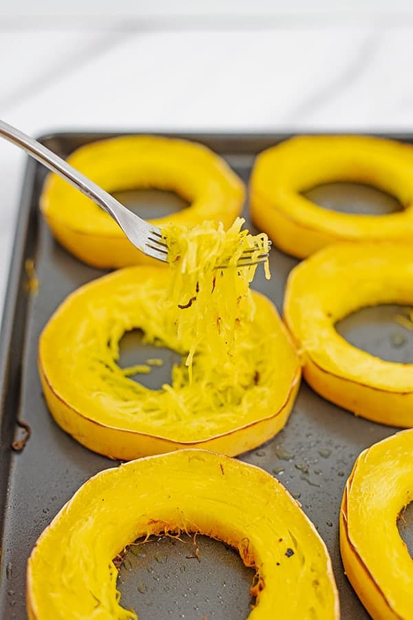 Baking sheet with spaghetti squash rings with a fork pulling strands of spaghetti squash from the rings.