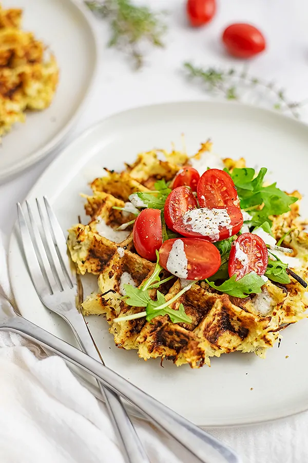 Close up of white plate with parsnip savory waffle recipe topped with arugula, tomatoes and dressing with two forks on the left side of the plate.