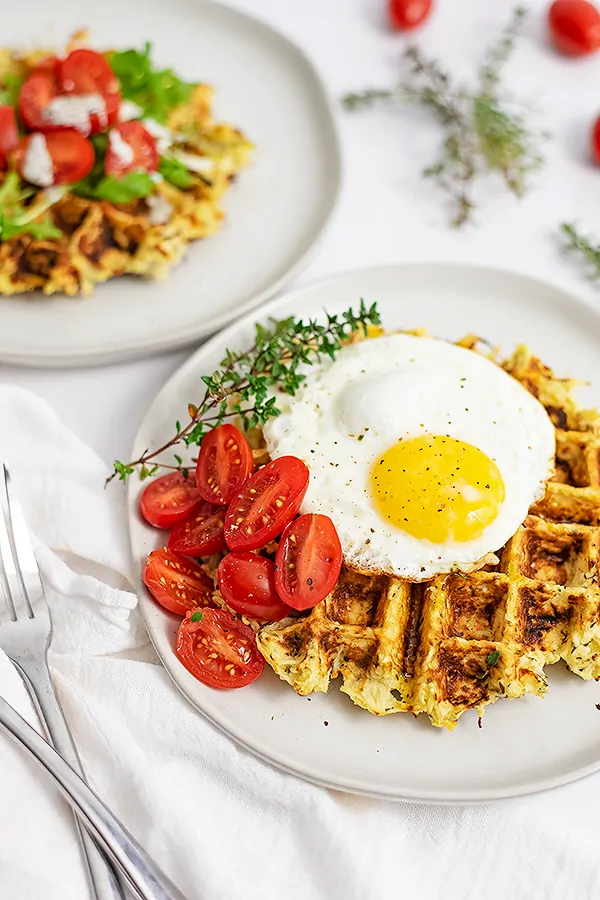 5 ingredient parsnip waffle savory waffle recipe on a white plate topped with a fried egg, tomatoes and sprigs of thyme. Second savory parsnip waffle in the background