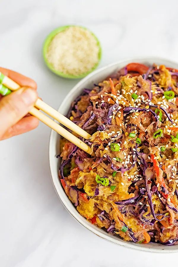 Hand holding chopsticks that are in a bowl of creamy Asian vegan spaghetti squash recipe with a small bowl of sesame seeds in the background