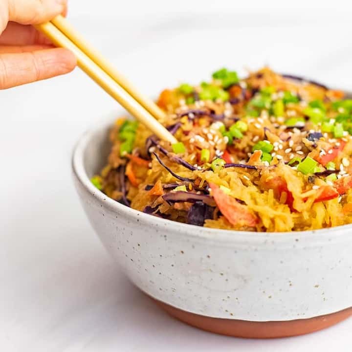 A large bowl with a rust colored bottom filled with creamy Asian vegan spaghetti squash recipe with chopsticks being held, plunging into the left side of the bowl