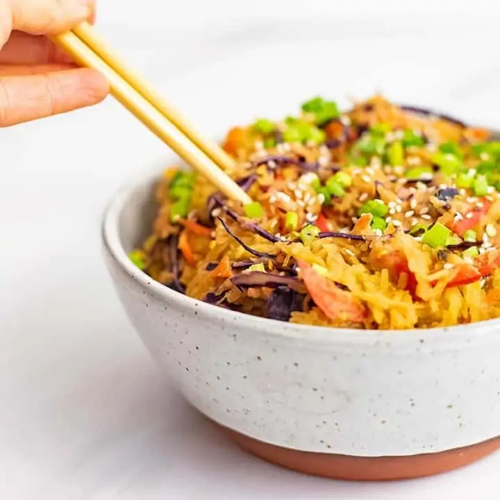 A large bowl with a rust colored bottom filled with creamy Asian vegan spaghetti squash recipe with chopsticks being held, plunging into the left side of the bowl