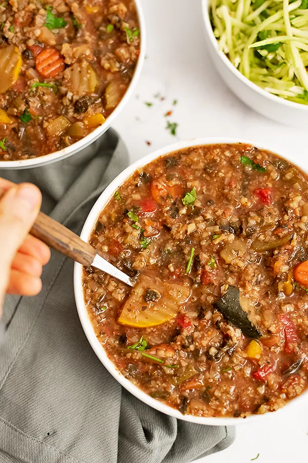 Hand holding a spoon over a bowl of crockpot low carb vegetable soup with a grey napkin next to the bowl