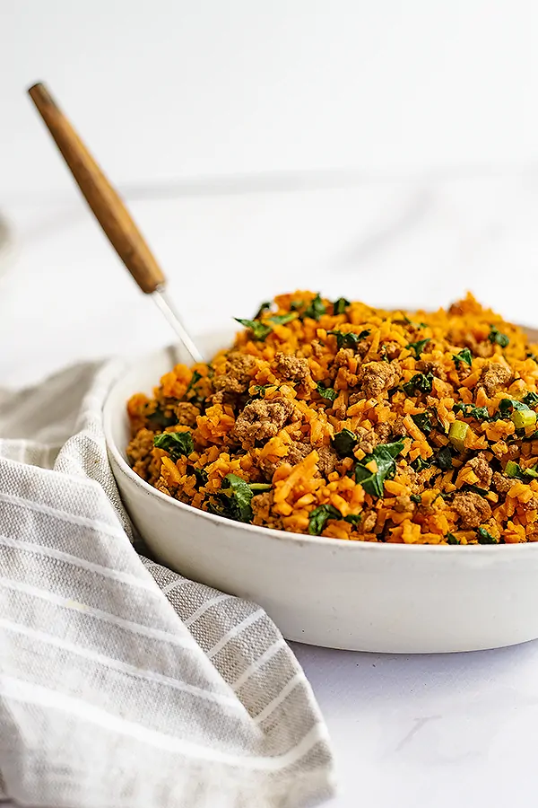 Large white bowl filled with the easy ground beef and sweet potato rice breakfast skillet recipe with a grey striped napkin to the left of the bowl and a wooden handled spoon in the bowl. 