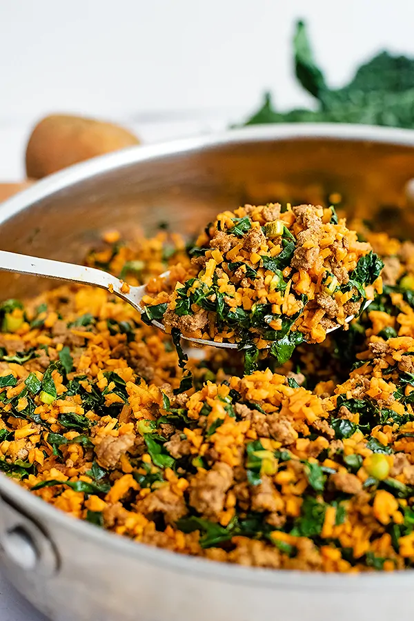 Large serving spoon scooping out a serving of the easy ground beef sweet potato rice breakfast skillet recipe with the skillet full of the recipe below the spoon