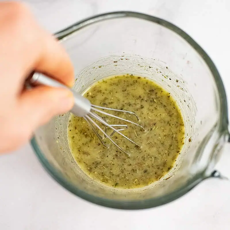 Hand holding a small whisk, stirring together lemon dressing in glass measuring cup. 