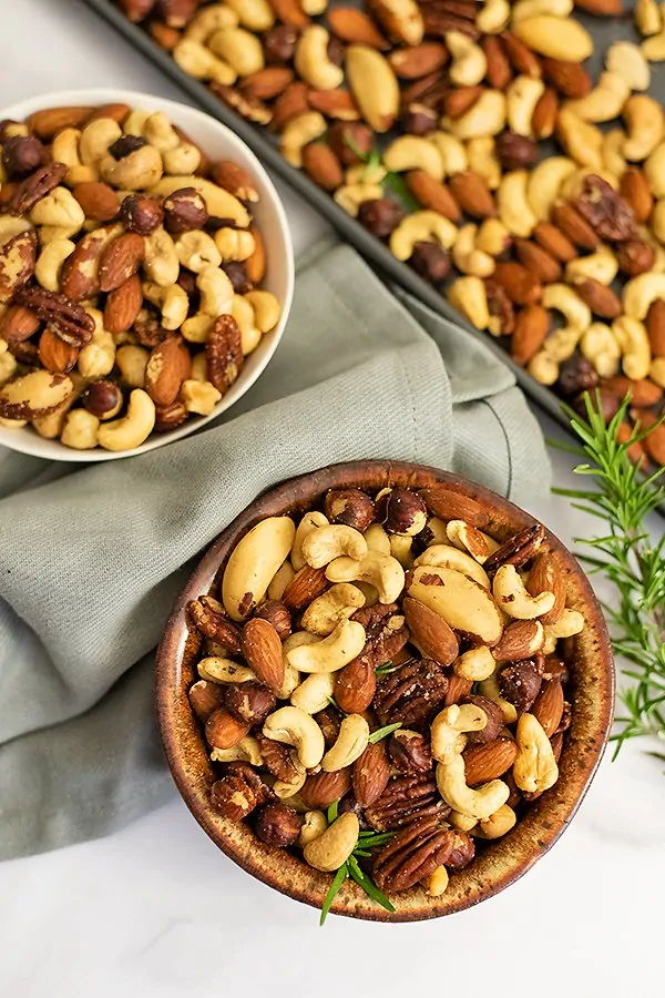 Overhead shot of two bowls of rosemary savory spiced nuts with a grey napkin between the bowls and a baking sheet full of nuts in the background
