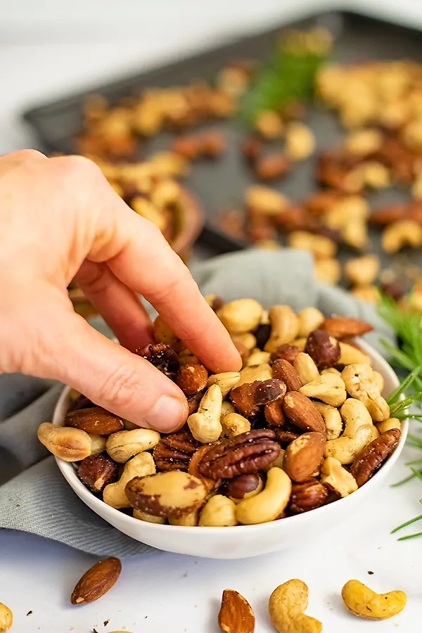 Hand reaching from the left into a white bowl filled with rosemary savory spiced nuts