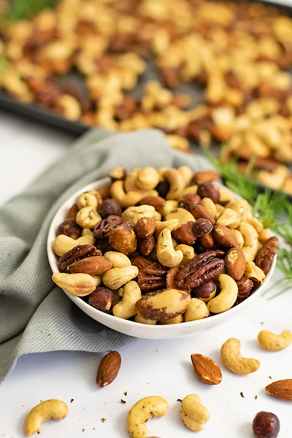 Close up on a white bowl filled with rosemary savory spiced nuts with a grey napkin and baking sheet full of nuts in the background