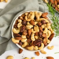 Overhead shot of white bowl filled with rosemary savory spiced nuts over a grey napkin with a rosemary sprig to the right of the bowl