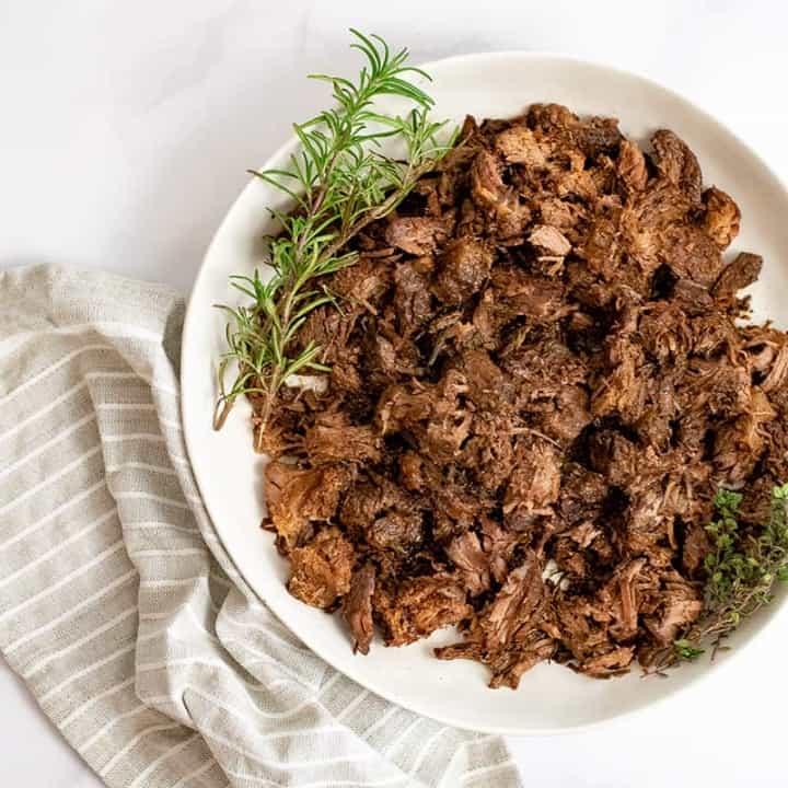 Overhead shot of large plate of slow cooker balsamic beef with fresh rosemary sprigs on the plate over a brown striped napkin