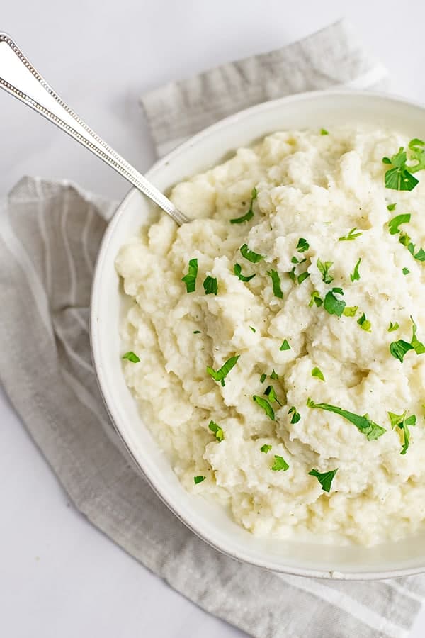 Overhead shot of a large bowl of vegan garlic mashed cauliflower over a grey striped napkin with a spoon in the bowl 