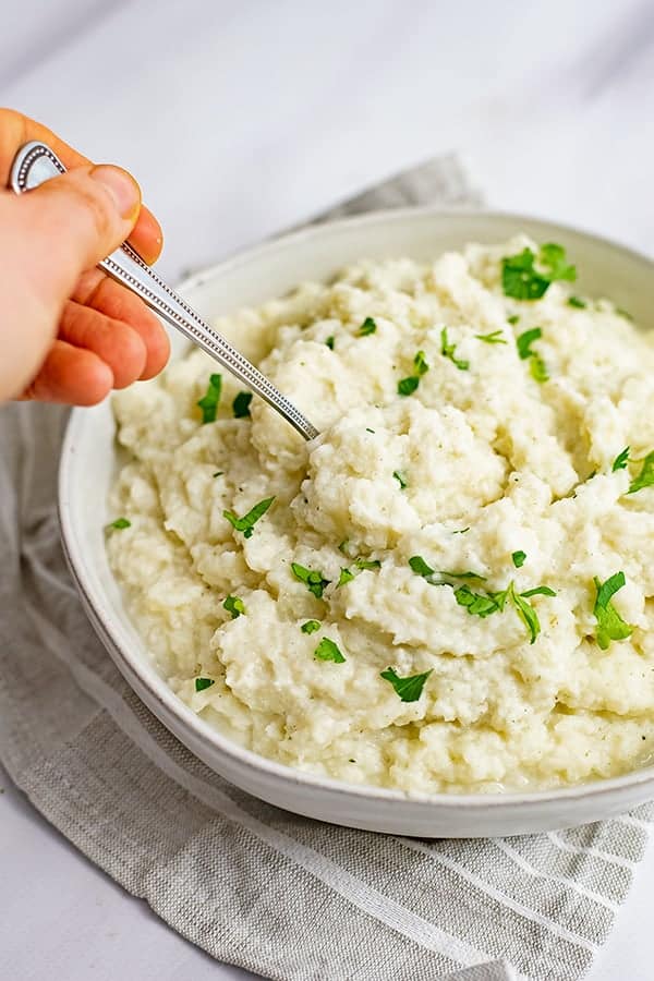 Overhead shot of a hand holding a spoon scooping out a serving of vegan garlic mashed cauliflower from a large bowl over a grey striped napkin