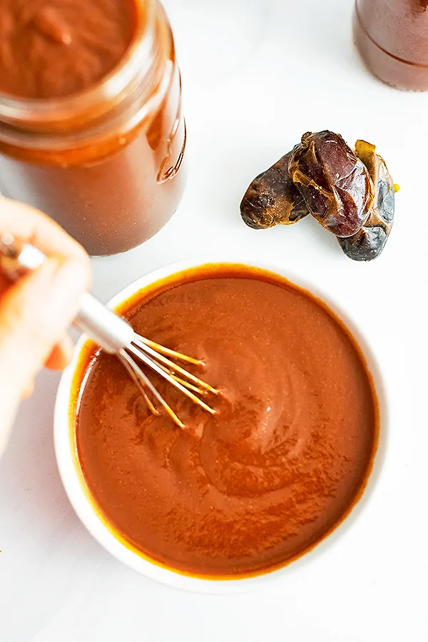 White bowl of Whole30 BBQ Sauce with a whisk stirring the sauce in the bowl. Dates and a jar of BBQ sauce in the background