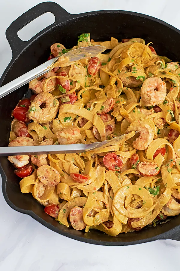 Cast iron skillet filled with Whole30 Creamy Cajun Shrimp Pasta recipe with a pair of silver tongs in the skillet