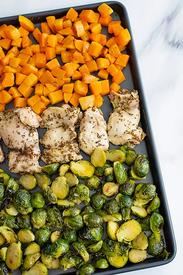 Sheet pan filled with butternuts squash cubes, dijon mustard chicken thighs and halved brussel sprouts. Pan is slightly off center