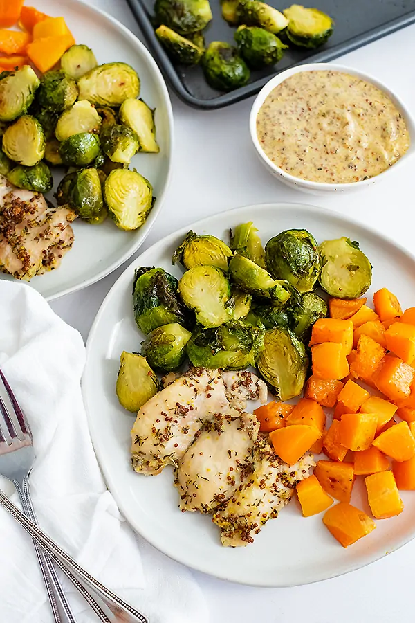 Sheet pan dijon mustard chicken thighs and vegetables plated on a white plate with a white napkin on the left of the plate and a dipping sauce in the background