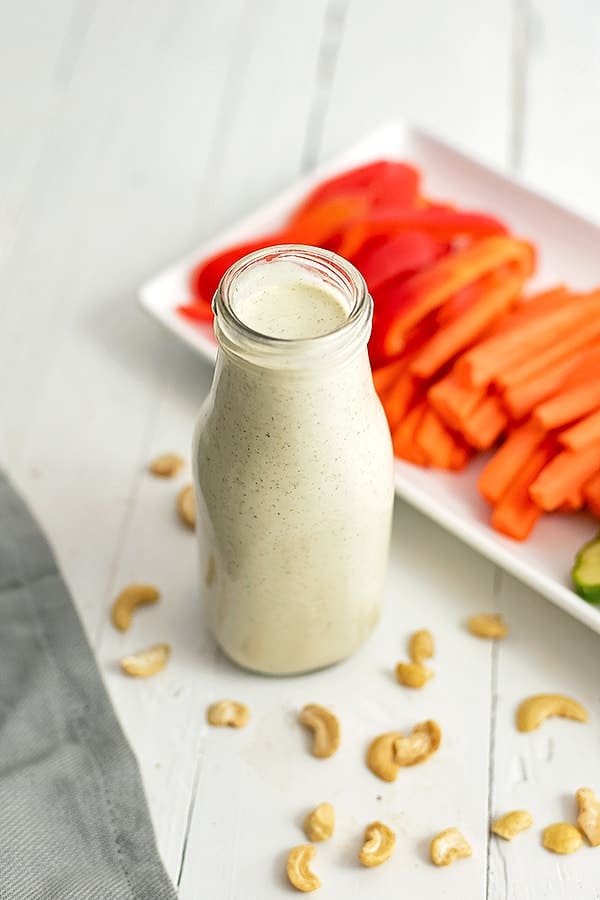 Bottle of dairy free ranch dressing with a platter of veggies in the background and cashews around the bottle on the table