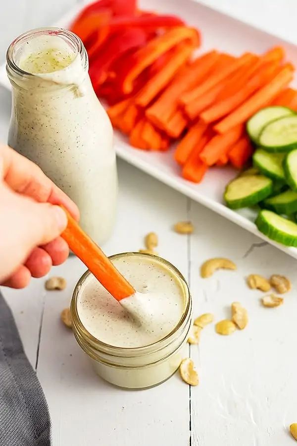 Small jar of dairy free ranch dressing with a carrot being dipped into the dressing. A bottle of dressing and a plate of veggies in the background