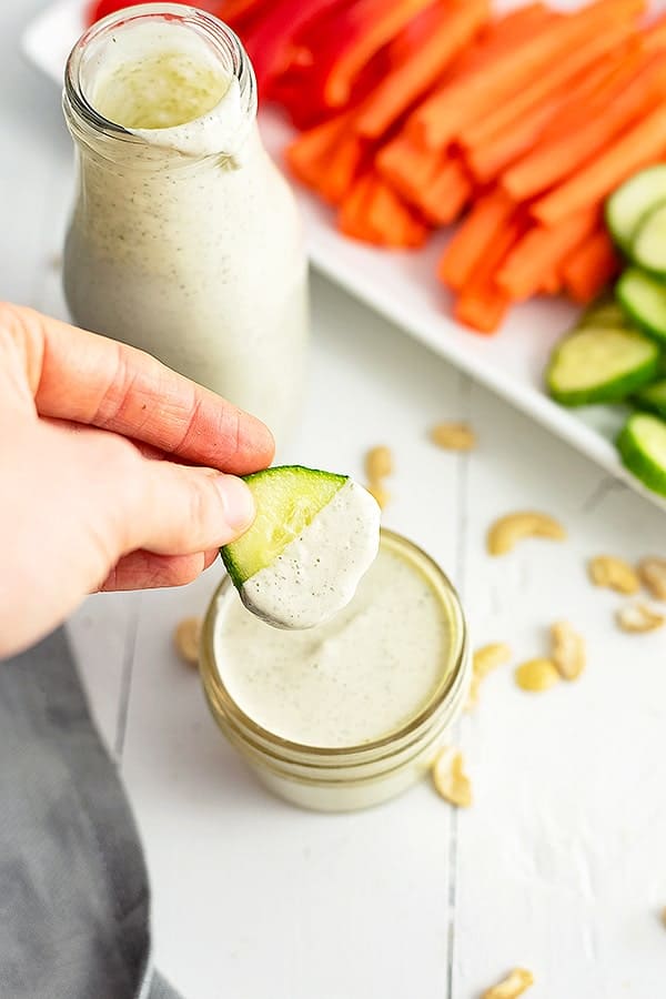Small jar of dairy free ranch dressing with a cucumber slice being dipped into the dressing. A bottle of dressing and a plate of veggies in the background
