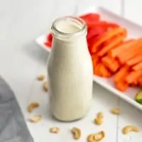 Bottle of dairy free ranch dressing on a white table with cashews on the table around the bottle. Platter of veggies in the background